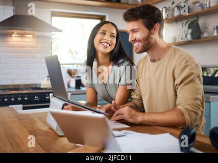 Happy young caucasian man working on laptop in the kitchen at home while his wife stands next to him and smiles while he looks at screen. Happy young Stock Photo