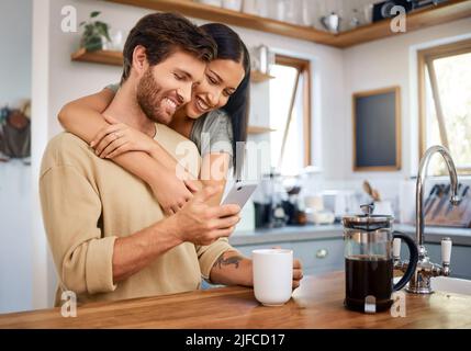 Happy young interracial couple being loving and affectionate at home. Young man using his smartphone and holds coffee cup while his girlfriend Stock Photo