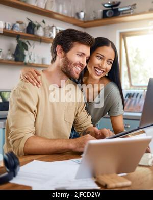 Beautiful young wife standing behind her husband working on his laptop while they look at the screen together and smile. Happy young interracial Stock Photo