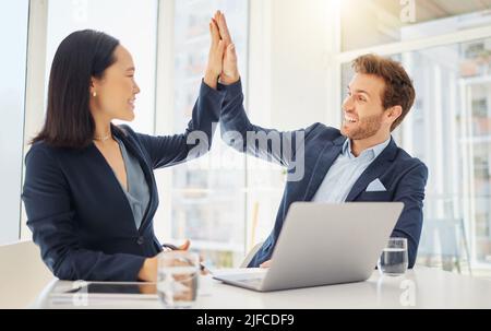 Two happy young diverse colleagues giving each other a high five while working together on a laptop in an office. Excited caucasian businessman Stock Photo