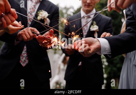 A crowd of young happy people in suits with sparklers in their hands during celebration. Sparkler on a wedding - bride, groom and guests holding light Stock Photo