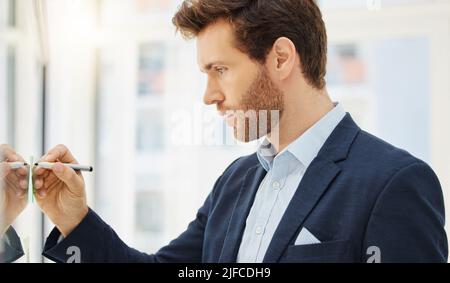One focused young caucasian businessman writing ideas on a glass wall while planning a project in an office. Ambitious entrepreneur brainstorming a Stock Photo