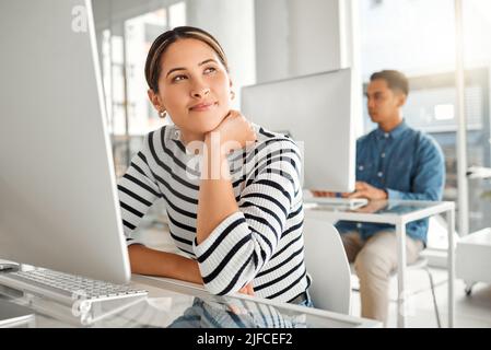 Young content mixed race businesswoman thinking and using a desktop computer in an office at work. Happy hispanic female businessperson smiling while Stock Photo