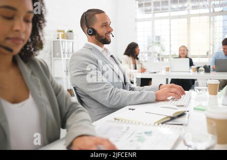 Young cheerful mixed race male call center agent answering calls while wearing a headset at work. Hispanic businessman talking on a call while typing Stock Photo