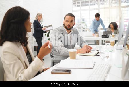 Young diverse colleagues talking while working on desktop computers together at a desk in an office at work. Young mixed race businessman talking to Stock Photo