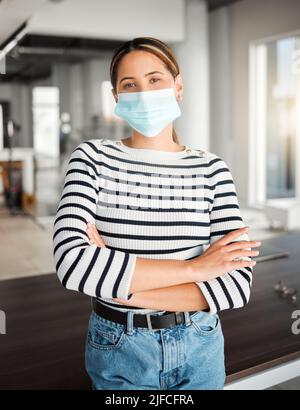 Young mixed race businesswoman standing with her arms crossed and wearing a mask at work. One hispanic female boss wearing a mask protecting from a Stock Photo