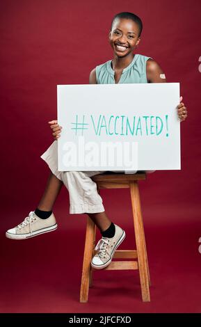 African american covid vaccinated woman showing and holding poster. Portrait of smiling black woman isolated against red studio background with Stock Photo