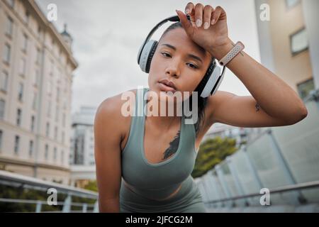 Portrait of a young mixed race female athlete wearing wireless headphones and listening to music while standing outside in the city wiping her sweaty Stock Photo