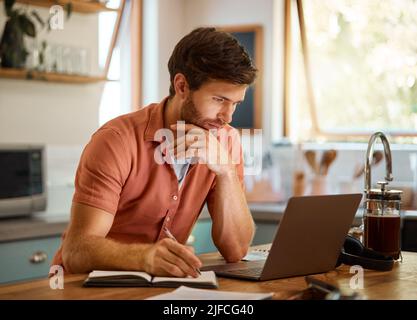 Young caucasian businessman writing in a notebook and working on a laptop at home alone. Focused male businessperson taking notes in a diary while Stock Photo