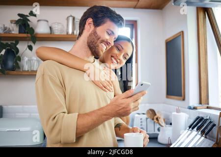 Happy young interracial couple being loving and affectionate at home. Young man using his smartphone and holds coffee cup while his girlfriend Stock Photo