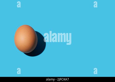 A brown chicken egg lies on the left on a bright blue background. Bright light, deep shadow. Close-up, copy space, top view. Stock Photo
