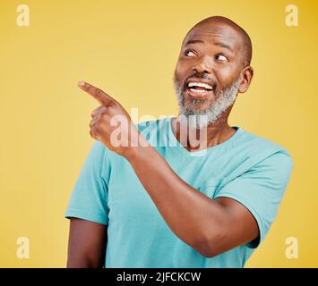 Mature african man smiling and pointing in a direction against a yellow studio background. Black guy looking happy and making a pointing gesture Stock Photo
