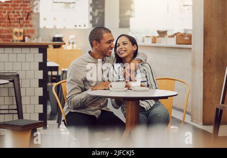 Loving young man sitting with his arm around his girlfriend while sitting at a table and having coffee in a cafe. Loving mixed race couple talking and Stock Photo