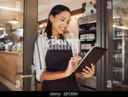 Businesswoman using a digital tablet in her restaurant. Cafe owner using an online app in her shop entrance. Confident business owner using a wireless Stock Photo