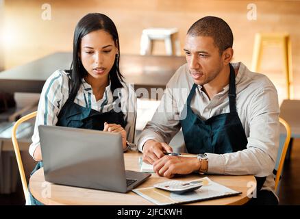 Two young mixed race employees wearing aprons sitting at a table in a coffee shop using a laptop and calculator at work. Young male and female Stock Photo