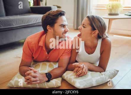 Funny romantic caucasian couple being playful pulling faces at each other while lying on the living room floor. Young man and woman looking at each Stock Photo