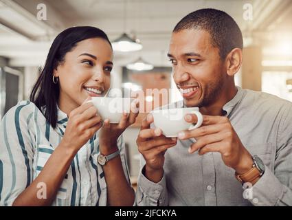 Loving mixed race couple in love looking at each other while holding cups enjoying coffee in a cafe. Happy young woman and man on their first date in Stock Photo