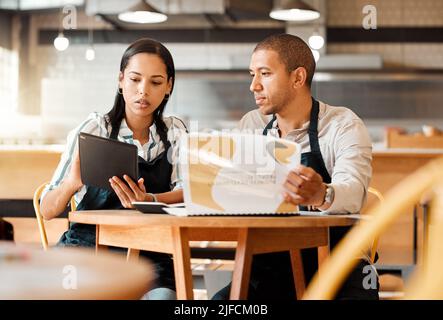 Two restaurant workers planning on a digital tablet in a small business. Young mixed race colleagues reviewing plans and reports in a cafe. Colleagues Stock Photo