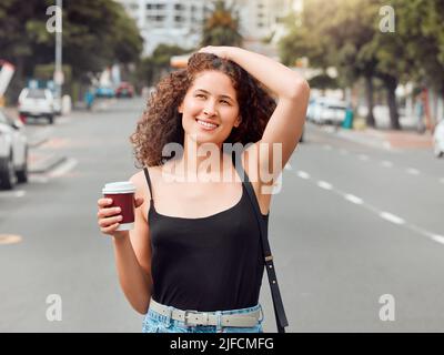 One happy beautiful young mixed race woman exploring the city while holding a takeaway coffee and touching her curly brunette hair. Hispanic Stock Photo