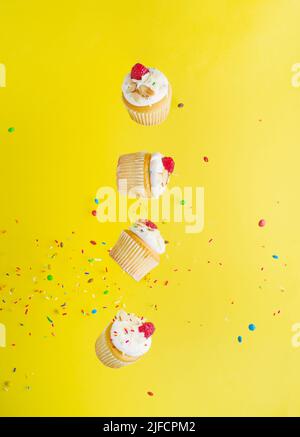 On a bright yellow background with colorful confetti, small cupcakes with cream and raspberries in a frozen flight. Festive composition. Children's an Stock Photo