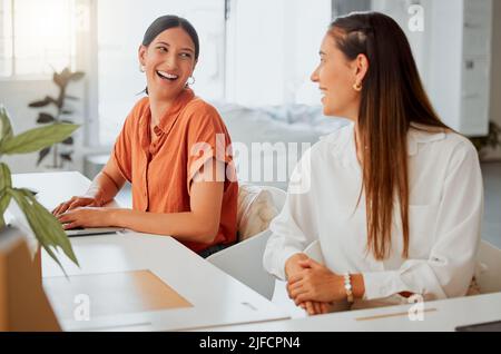 Cheerful young hispanic business woman working on a laptop while talking to a colleague in an office. Two happy coworkers working together in a Stock Photo