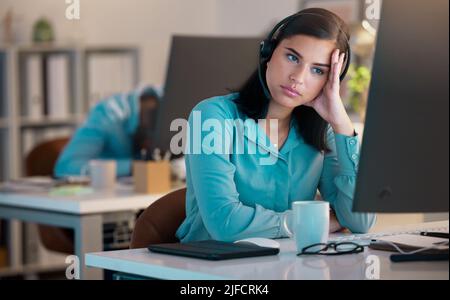 One stressed young caucasian call centre telemarketing agent looking bored with headache while working on computer in an office. Female consultant Stock Photo