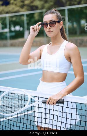 Trendy young tennis player wearing glasses and holding tennis racket while leaning on a net. Confident young sportswoman ready for a tennis match Stock Photo