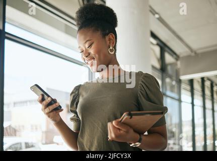 African american business woman smiling while reading or texting on smartphone holding journal and walking in modern office. Smiling female Stock Photo