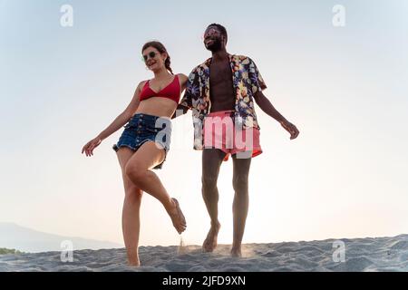 Engaged biracial couple happily walking barefoot in the sand holding together - multi-ethnic people relationship in the summer lifestyle concept Stock Photo