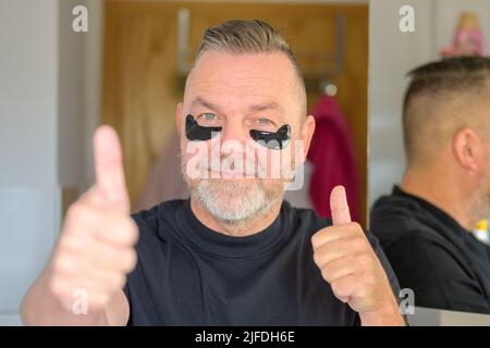 Senior man using gel hydrating eye patches giving a double thumbs up of approval in a skin care concept Stock Photo