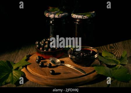 Blackcurrant in a ceramic bowl and blackcurrant jam in a glass bowl on a wooden board with green currant leaves and glass jars of jam in the backgroun Stock Photo