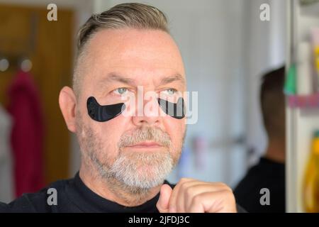 Middle-aged man applying gel hydration patches to the pouches below his eyes in a male skincare concept Stock Photo