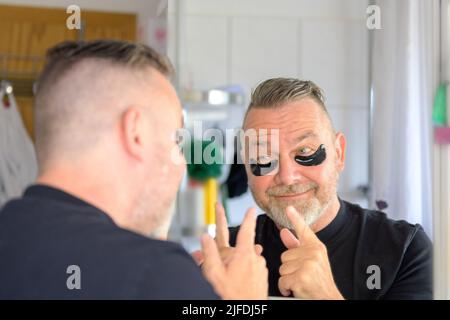 Man using hydrating gel eye patches pointing to himself in the bathroom mirror with a smile in a skin care concept Stock Photo