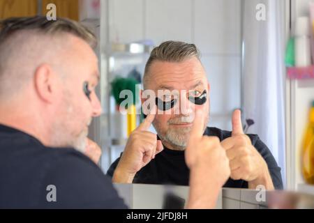 Senior man pointing to hydrating gel eye patches while giving a thumbs up gesture viewed over the shoulder to his reflection in the mirror Stock Photo