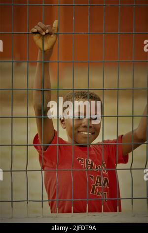 A vertical shot of a happy kid wearing a red shirt behind the fence in a blurred background Stock Photo