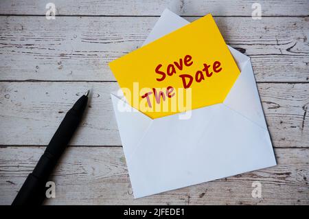 Save the date text on yellow notepad in an envelope with pen and wooden desk background. Stock Photo