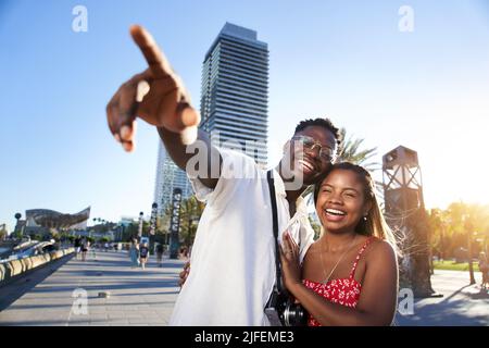 Smiling young African-American traveling couple enjoy a summer vacation in a resort town. The boyfriend points out something on the horizon to his Stock Photo
