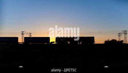DB cargo Rail UK class 66 diesel locomotive making a sunset silhouette with a container freight train on the electrified west coast mainline Stock Photo