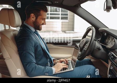 Waist up side view portrait of happy elegant male sitting in salon of his car while typing something on keyboard of laptop Stock Photo