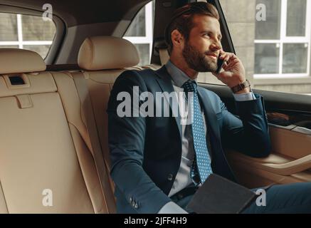 Waist up side view portrait of handsome bearded boss in formal wear sitting on back seat of a car while talking on mobile phone Stock Photo