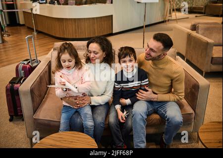 Adorable loving family- mom and dad, gently hugging their kids playing with toy planes while waiting the flight boarding in the VIP lounge area of the Stock Photo