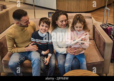 Happy multi-ethnic young loving mom and dad spending time together playing toy planes with their kids, son and daughter at the airport while waiting f Stock Photo