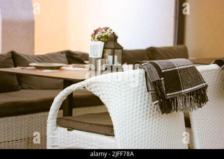 White chair on a summer patio, terrace cafe, restaurant. Checkered brown plaid on a back of a stool. Comfortable rattan garden furniture. Decorative l Stock Photo