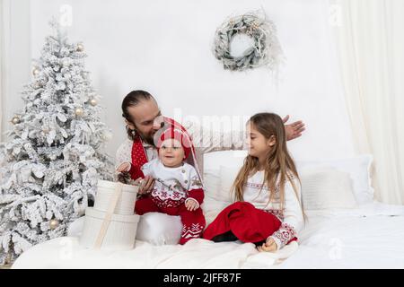 Kid sits on dad's lap and enjoys Christmas morning with his sister opening presents. Concept of happy holidays with children and parents. Front view. Stock Photo