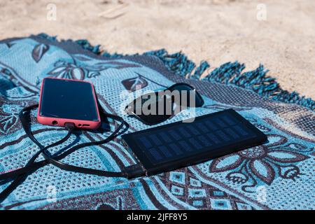 Solar panel charging a mobile phone on a beach. Portable powerbank solar charger in sunlight. Sun energy. Stock Photo
