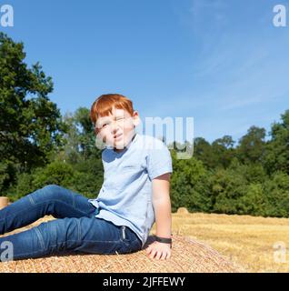 a red-haired boy sits on top of a golden straw stack in a field, a boy on a prickly wheat straw stack, a portrait of a seven-year-old boy against a bl Stock Photo