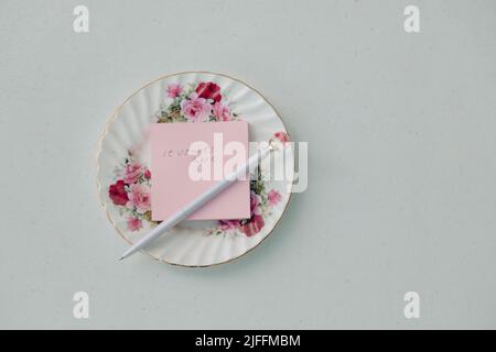 Pink sticky note on floral plate with rose pen, text states 'It starts here...' Flat lay image Stock Photo