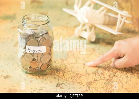 Hand pointing attractions in a map on a table with a glass jar with coins and travel inscriptions and a wooden toy plane nearby. Stock Photo