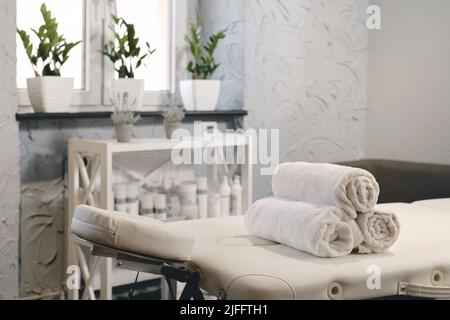 Rolled white towels on massage table in empty salon. Interior of aesthetic clinic, nobody. Modern cabinet of physiotherapist. Empty room with plants Stock Photo