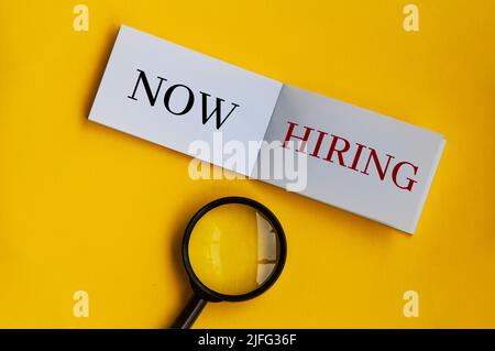 Now hiring text on notepad with magnifying glass on yellow background. Employment concept Stock Photo